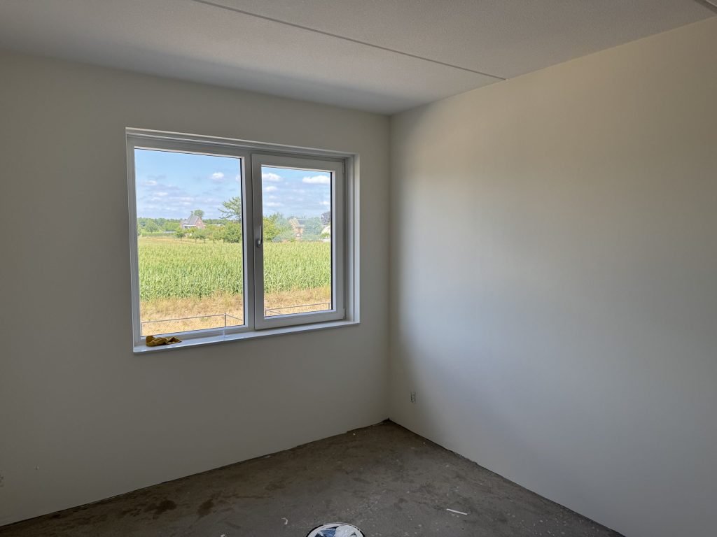 Kamer met strak afgewerkte lichte wanden en een raam met uitzicht op een maisveld